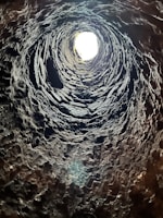A vertical view from the bottom of a deep, rugged, stone-lined well or shaft with a bright light at the opening. The rough textures of the stone are prominent, and shadows create a spiral effect as they lead upwards towards the light source.