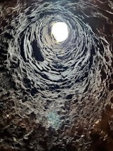 A vertical view from the bottom of a deep, rugged, stone-lined well or shaft with a bright light at the opening. The rough textures of the stone are prominent, and shadows create a spiral effect as they lead upwards towards the light source.
