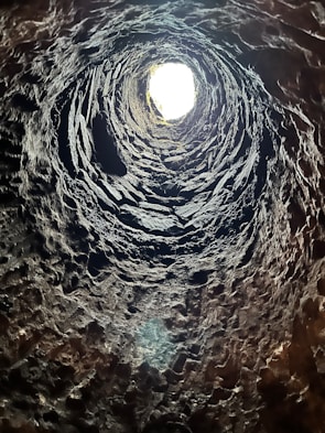 A vertical view from the bottom of a deep, rugged, stone-lined well or shaft with a bright light at the opening. The rough textures of the stone are prominent, and shadows create a spiral effect as they lead upwards towards the light source.