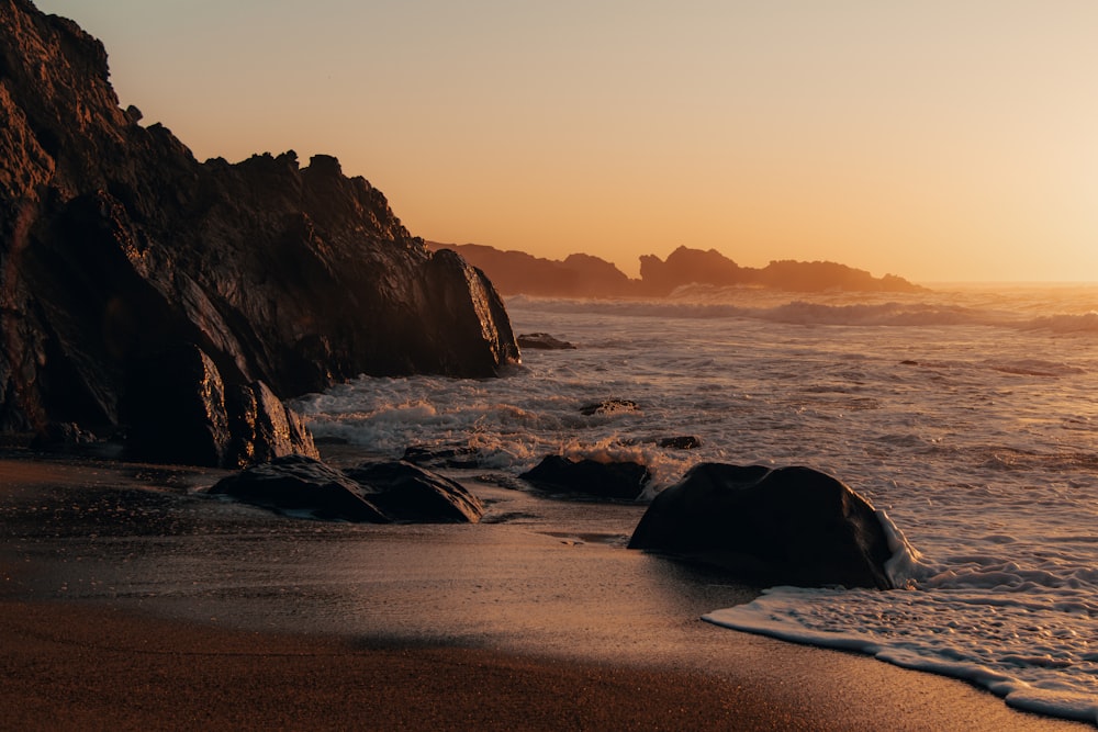 the sun is setting over the ocean with rocks in the foreground