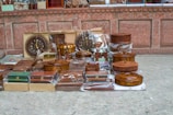A collection of intricately carved wooden items, including boxes, trays, and decorative pieces, is displayed on the ground against a rustic stone wall. Many items are encased in plastic, indicating their new or preserved condition. The display includes round, rectangular, and ornate pieces, some featuring intricate designs and craftsmanship.