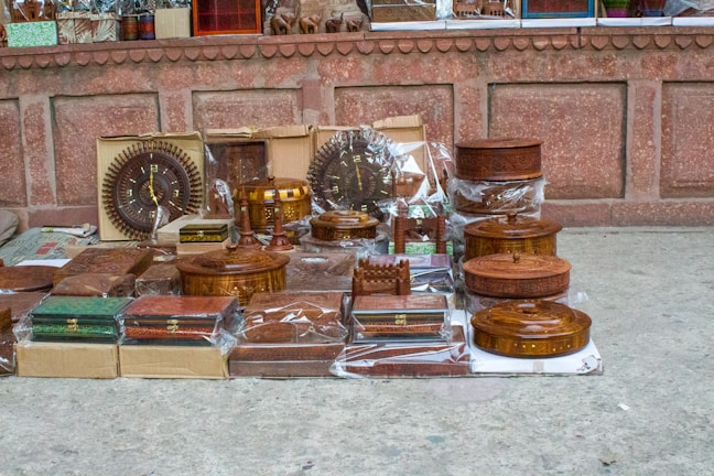 A collection of intricately carved wooden items, including boxes, trays, and decorative pieces, is displayed on the ground against a rustic stone wall. Many items are encased in plastic, indicating their new or preserved condition. The display includes round, rectangular, and ornate pieces, some featuring intricate designs and craftsmanship.