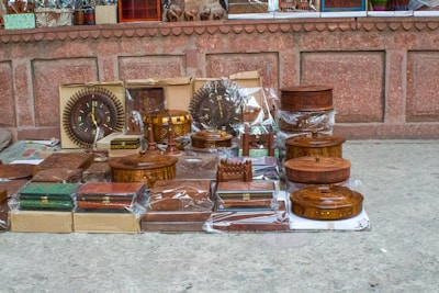 A collection of intricately carved wooden items, including boxes, trays, and decorative pieces, is displayed on the ground against a rustic stone wall. Many items are encased in plastic, indicating their new or preserved condition. The display includes round, rectangular, and ornate pieces, some featuring intricate designs and craftsmanship.