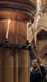 A person is lighting candles on a tall, ornate candle holder attached to a stone pillar inside a cathedral or church. Smoke rises from the lit candles, and the architecture features arched ceilings and intricate stonework.