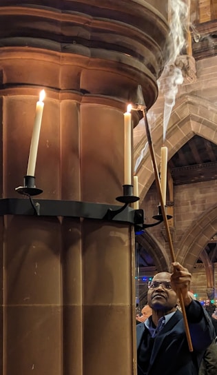 A person is lighting candles on a tall, ornate candle holder attached to a stone pillar inside a cathedral or church. Smoke rises from the lit candles, and the architecture features arched ceilings and intricate stonework.