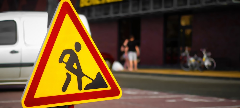 A triangular warning sign displayed prominently, indicating road work. The sign features a black silhouette of a person working with a shovel against a bright yellow background with a red border. In the blurred background, there is a white van and a group of people along with some bicycles.