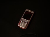 A classic mobile phone with a numeric keypad and small screen rests on a dark surface. The phone has a metallic finish with a black display and visible branding near the earpiece.