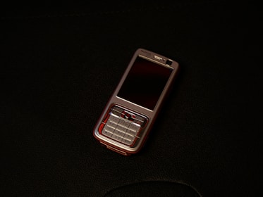 A classic mobile phone with a numeric keypad and small screen rests on a dark surface. The phone has a metallic finish with a black display and visible branding near the earpiece.