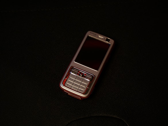 A classic mobile phone with a numeric keypad and small screen rests on a dark surface. The phone has a metallic finish with a black display and visible branding near the earpiece.