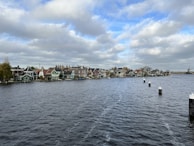 A picturesque row of traditional Dutch houses lines the edge of a wide river, with a faint outline of a windmill visible in the distance on the right. The sky is partially cloudy, casting a soft light on the scene, and the water in the foreground is gently textured by waves.