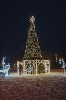 A large, beautifully decorated Christmas tree is adorned with twinkling lights and topped with a bright star. Surrounding the tree are illuminated figures and decorative arches, set against a snowy ground. The scene is enhanced by a subtle snowfall, adding to the festive atmosphere.