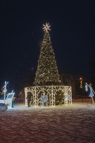 A large, beautifully decorated Christmas tree is adorned with twinkling lights and topped with a bright star. Surrounding the tree are illuminated figures and decorative arches, set against a snowy ground. The scene is enhanced by a subtle snowfall, adding to the festive atmosphere.
