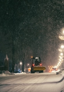 A snow-covered street being plowed by a large snowplow.