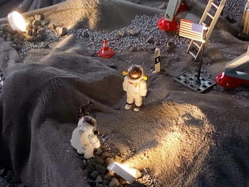 A miniature lunar scene with two astronaut figurines in white spacesuits exploring a rocky, gray surface. The setup is illuminated by a light, casting shadows across the terrain. A small American flag is displayed on the right, next to a ladder that is part of what looks like a lunar module replica. Several small rocks and a traffic cone are scattered across the surface.