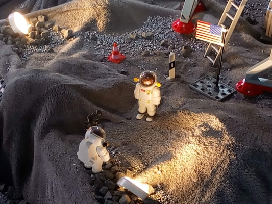 A miniature lunar scene with two astronaut figurines in white spacesuits exploring a rocky, gray surface. The setup is illuminated by a light, casting shadows across the terrain. A small American flag is displayed on the right, next to a ladder that is part of what looks like a lunar module replica. Several small rocks and a traffic cone are scattered across the surface.
