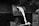 Molten metal is being poured from a furnace. A worker's gloved hand holds the handle of a container, directing the bright, flowing liquid into a mold below. The surrounding environment is industrial, with a focus on the intense heat and fluid motion of the metal.