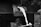 Molten metal is being poured from a furnace. A worker's gloved hand holds the handle of a container, directing the bright, flowing liquid into a mold below. The surrounding environment is industrial, with a focus on the intense heat and fluid motion of the metal.