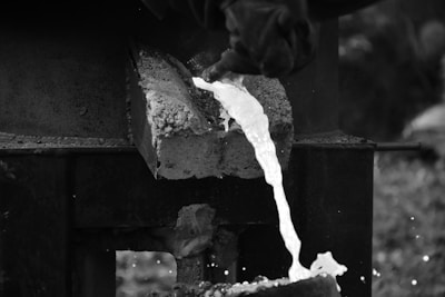 Molten metal is being poured from a furnace. A worker's gloved hand holds the handle of a container, directing the bright, flowing liquid into a mold below. The surrounding environment is industrial, with a focus on the intense heat and fluid motion of the metal.