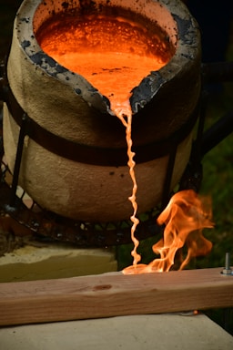 Molten metal flows from a crucible, creating a glowing, orange stream. Flames rise up as the hot metal makes contact with a wooden surface. The crucible appears to be heat-stained and is positioned on a support structure, with the whole setup situated outdoors.