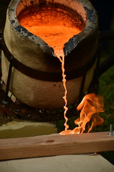 Molten metal flows from a crucible, creating a glowing, orange stream. Flames rise up as the hot metal makes contact with a wooden surface. The crucible appears to be heat-stained and is positioned on a support structure, with the whole setup situated outdoors.