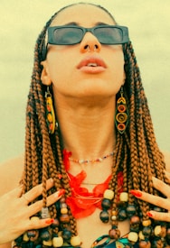A person wearing a collection of colorful jewelry with intricate braided hair, adorned with beads. The individual has sunglasses and large, vibrant earrings resembling spiral designs. The hands are positioned near the neck, showing painted nails and multiple rings.