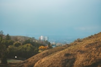 A landscape view featuring a city in the background, surrounded by hills and greenery. In the distance, a white building is visible amid a hazy sky. In the foreground, there are dry, brown hills, with patches of green trees and a basketball court.