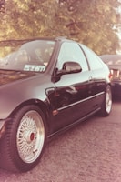 A black car parked on a road, featuring custom rims with a vintage design. A paper with numbers is visible on the windshield. The scene includes another vehicle partially visible in the background, with trees overhead.