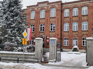 A red brick building with symmetrical windows and an 'AD 1903' plaque is visible. The front entrance is bordered by concrete pillars and an iron gate, with snow covering the ground and a large pine tree to the left. A Polish flag and road signs are situated near the entrance.