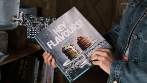 A person in a denim jacket holds a cookbook titled 'HEY FLAVOURS!' featuring a decadent image of a stack of pancakes topped with berries and syrup on its cover. The book appears to be on a shelf next to a vintage water dispenser and some stacked glass cups. Additional books with Japanese text are also visible in the background.