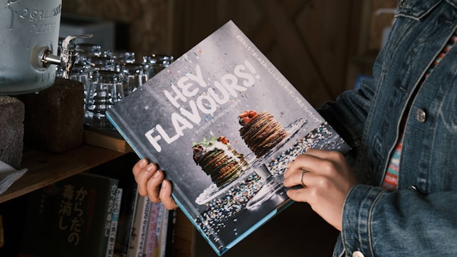 A person in a denim jacket holds a cookbook titled 'HEY FLAVOURS!' featuring a decadent image of a stack of pancakes topped with berries and syrup on its cover. The book appears to be on a shelf next to a vintage water dispenser and some stacked glass cups. Additional books with Japanese text are also visible in the background.