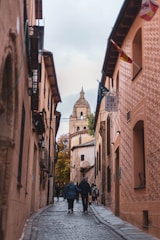 A narrow, cobblestone street lined with old, rustic buildings leads toward a distant historical tower with a dome. Flags hang from the buildings, and groups of people can be seen walking along the street. The sky overhead is overcast, and trees with autumnal leaves are visible.