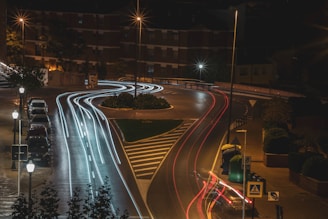 Long exposure photograph of a street at night with light trails from moving vehicles. Streetlamps illuminate the road, lined with parked cars on one side. Buildings and trees surround the area, adding to the urban atmosphere.