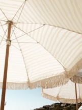Large outdoor umbrellas with fringe trim open against a clear blue sky. The fabric has a light beige and white striped pattern. In the distance, there is a view of some greenery and hilltop buildings.