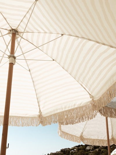 Large outdoor umbrellas with fringe trim open against a clear blue sky. The fabric has a light beige and white striped pattern. In the distance, there is a view of some greenery and hilltop buildings.