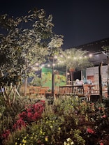 A cozy outdoor seating area featuring wooden benches surrounded by lush greenery and vibrant flowers. String lights hang above, illuminating the space at night, creating a warm and inviting atmosphere. A green wall with hanging potted plants adds to the garden-like setting.