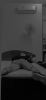 A person is lying face down on a bed with legs extended. An air conditioning unit is mounted on the wall above, and a fan is also visible. The room has a calm and minimalistic setting.