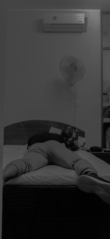 A person is lying face down on a bed with legs extended. An air conditioning unit is mounted on the wall above, and a fan is also visible. The room has a calm and minimalistic setting.