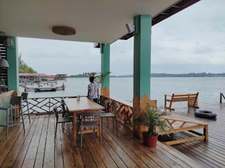 A wooden deck area with tables and chairs overlooking a calm body of water. In the foreground, a person is standing near a table, facing the water. The deck is partially covered by a roof supported by green pillars. There are potted plants and a few structures like a bench and a tire float on the deck. In the background, there are wooden buildings on stilts along the water, and the sky is overcast.