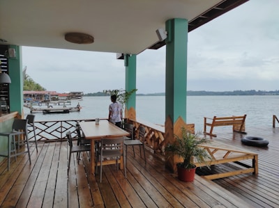 A wooden deck area with tables and chairs overlooking a calm body of water. In the foreground, a person is standing near a table, facing the water. The deck is partially covered by a roof supported by green pillars. There are potted plants and a few structures like a bench and a tire float on the deck. In the background, there are wooden buildings on stilts along the water, and the sky is overcast.