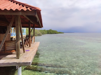 A wooden deck with a red-tiled roof extends over clear, shallow water, supported by wooden beams. Several wooden benches are placed under the shelter. The water is transparent, revealing the rocky bottom, and stretches into the horizon where it meets a cloudy sky. On the left, there are hints of bright blue structures, possibly a building or home, and lush green vegetation lines the distant shoreline.