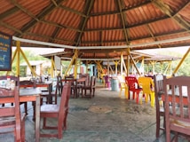 A rustic open-air dining area with a wooden roof structure, featuring a mix of wooden and plastic chairs, and tables that are neatly arranged. Brightly colored chairs in yellow and red add a lively touch to the setting. Small bottles and condiments are visible on the tables, indicating readiness for dining. Trees and greenery can be seen in the background, suggesting an outdoor or forest setting.