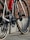 A close-up view of a road bicycle's rear section, showing gears, chain, and the red water bottle attached to the frame. The setting includes a brick wall backdrop and a stone-paved ground.