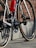 A close-up view of a road bicycle's rear section, showing gears, chain, and the red water bottle attached to the frame. The setting includes a brick wall backdrop and a stone-paved ground.