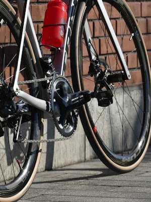 A close-up view of a road bicycle's rear section, showing gears, chain, and the red water bottle attached to the frame. The setting includes a brick wall backdrop and a stone-paved ground.