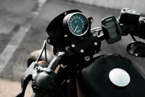 View of a motorcycle's handlebar and speedometer, with a focus on the round analog speed gauge and attached phone holder. The background is blurred, suggesting the motorcycle is stationary and parked on an asphalt surface.