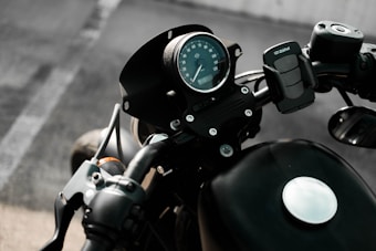 View of a motorcycle's handlebar and speedometer, with a focus on the round analog speed gauge and attached phone holder. The background is blurred, suggesting the motorcycle is stationary and parked on an asphalt surface.