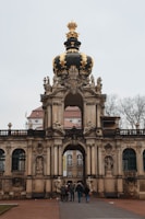 An ornate baroque gate features intricate sculptures and a prominent black and gold crown adorned on top. The structure stands tall with decorative architectural elements. Below, a group of people walks towards the entrance, with bare trees in the background adding to the historical ambiance.