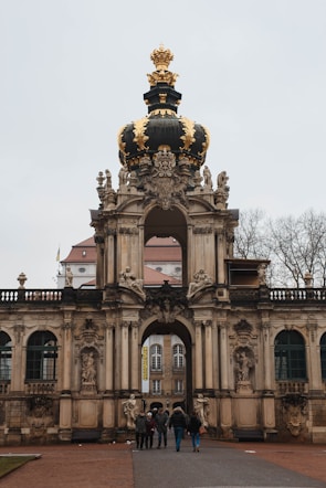An ornate baroque gate features intricate sculptures and a prominent black and gold crown adorned on top. The structure stands tall with decorative architectural elements. Below, a group of people walks towards the entrance, with bare trees in the background adding to the historical ambiance.