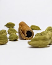 A cinnamon stick is surrounded by several cardamom pods, arranged on a white surface. The cinnamon is positioned centrally, with its textured, rolled shape creating a point of focus, while the cardamom pods are scattered around, adding depth and contrast.
