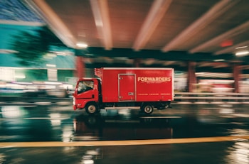 A red delivery truck is moving at high speed under an overpass on a wet road. The motion blur suggests fast movement, and the reflections on the pavement indicate recent rainfall. The truck has the word 'FORWARDERS' written on its side.